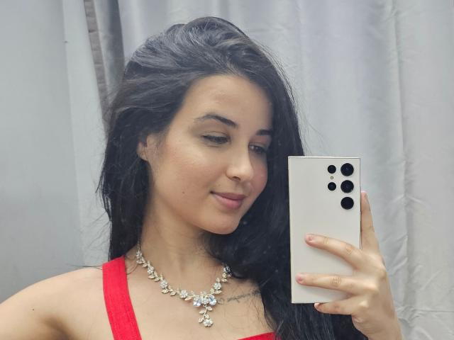 EsmeraldaXx with long black hair, red dress, and silver necklace, taking a selfie with a white iPhone, showing a subtle smile, holding the phone with her right hand.