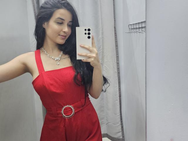 EsmeraldaXx with long black hair, wearing a red dress with a belt, holding a white phone, smiling, showing her tits, necklace, and looking at the mirror.