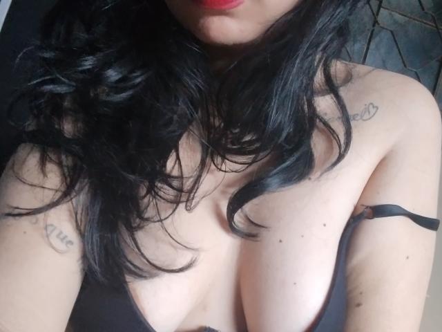 YosefinFabouls has black wavy hair, red lips, light skin, and large tits with tattoos on her shoulders, wearing a black strap dress, showing cleavage and a strap.