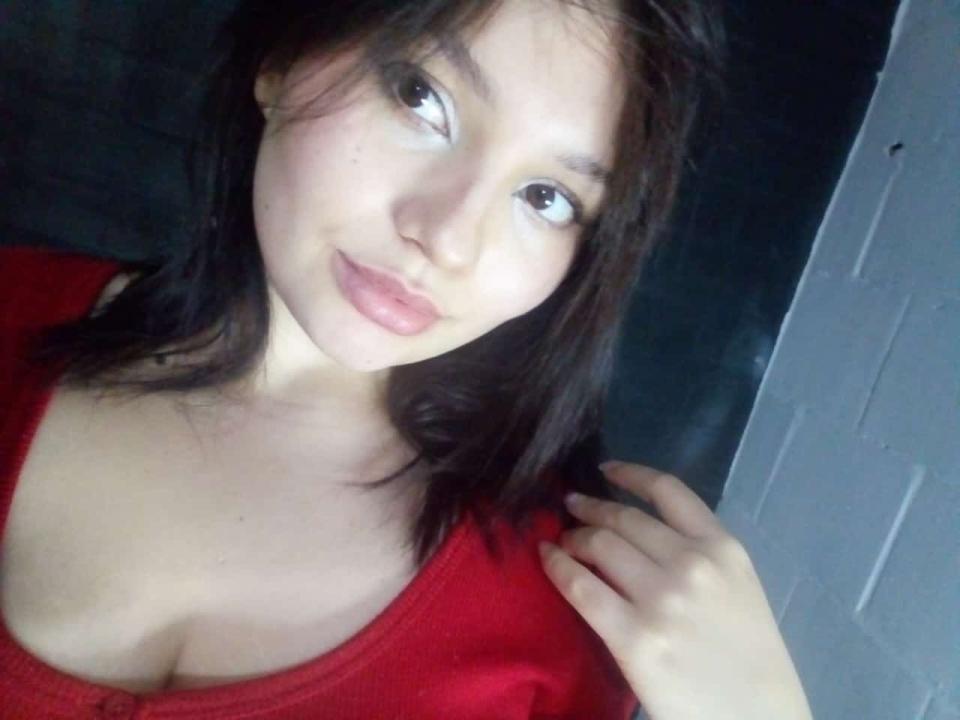 Starligth with light skin and dark brown hair, wearing a red shirt, showing her tits, looking up with a slight smile, hand near her shoulder, in a selfie.