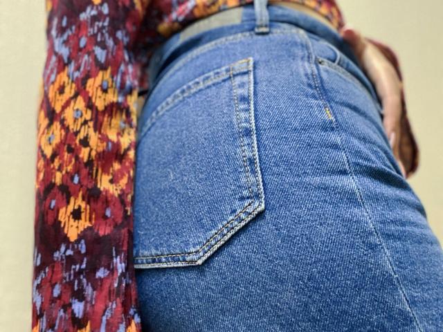Rubbi's blue denim butt with visible back pockets, wearing a colorful, patterned top. Her hand rests on her hip, showing a subtle tan line. Tight, textured jeans.