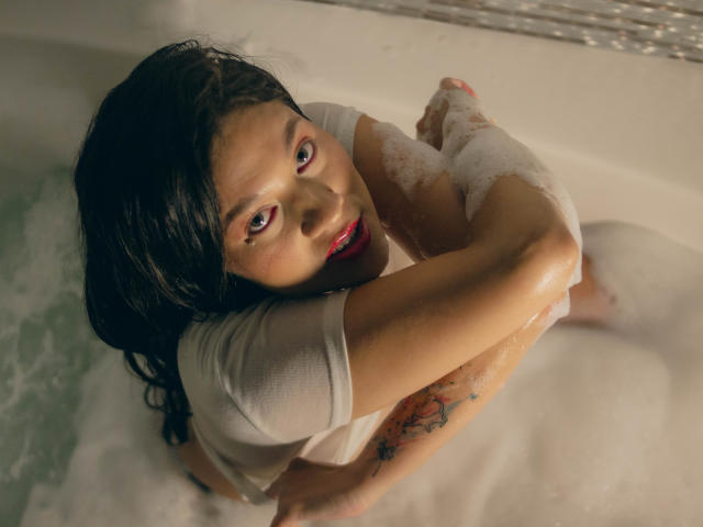 LindaJimenex in einer Badewanne mit schäumendem Wasser, Arme vor der Brust verschränkt, nasses weißes Hemd, dunkles, nasses Haar, roter Lippenstift, Tattoo am rechten Arm, blickt zur Kamera auf..