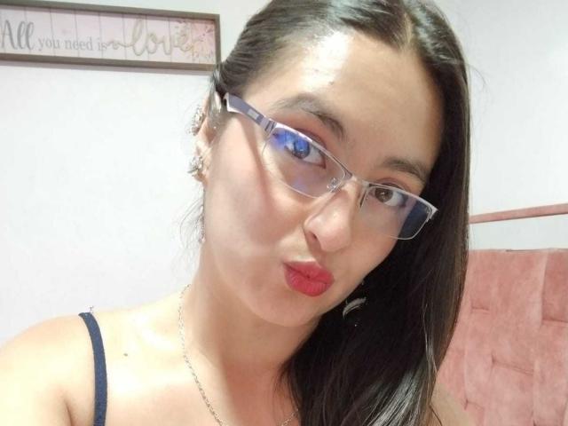 VenusSweetPalmer with long black hair, wearing glasses, red lipstick, and a black spaghetti strap top, pouting her full, shiny-assed lips. She has clear skin and a silver necklace.
