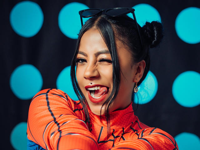 Amberv with dark hair in double buns, wearing an orange latex suit, has her mouth open, showing a small red lip piercing, and black sunglasses on her head.