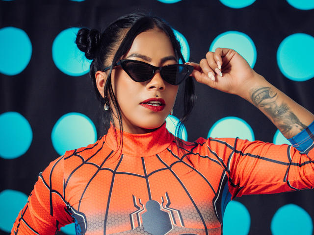 Amberv in a Spider-Man costume, wearing black sunglasses, adjusting them with her right hand, showing a tattoo on her forearm, with her black hair in a bun.