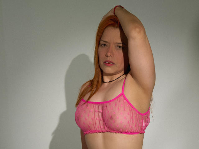 BiancaWild, con i capelli rossi, indossa un reggiseno trasparente rosa, alza il braccio destro, mostrando il seno, le labbra rosa, una collana nera e un leggero sorriso, su uno sfondo bianco uniforme..