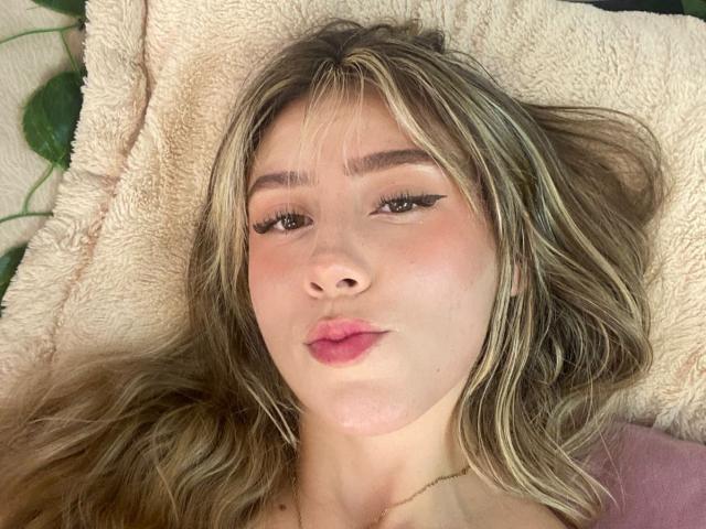 LivelyVanderWood's face, lying on a fluffy beige pillow, with long blonde hair, full pink lips, and winged eyeliner, looking directly at the camera. Her tits are partially visible.