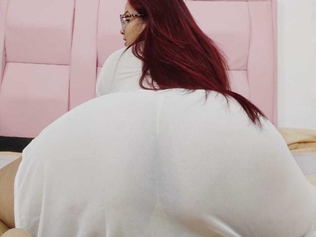 SophieTr's huge, white-covered ass is in the foreground, facing away. She has long, red hair and is wearing glasses. Her back is to the camera.
