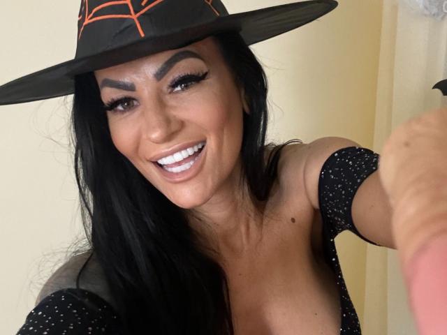 AndraD with black hair, wearing a witch hat, smiling, showing white teeth, and holding a phone. She's got a cute face, big boobs, and a black sparkly top.