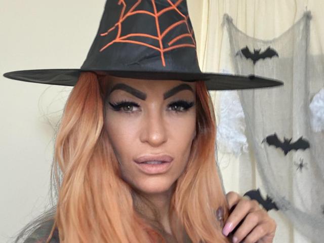 AndraD with long orange hair, black witch hat, orange spider web, and thick eyebrows, showing her pouty lips and dark eyeliner, has a sexy, cute look, with her hand up.