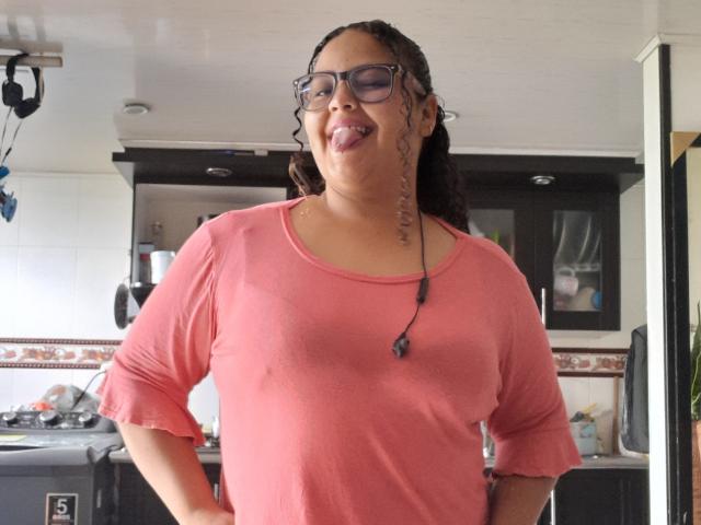 MelannieCarter with curly brown hair, glasses, and a pink long-sleeve shirt, sticking out her tongue, showing her small, round tits through the shirt. Black necklace with beads.