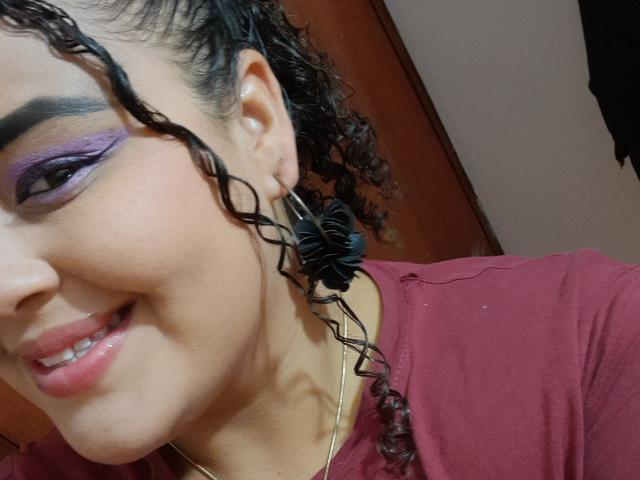 Melannie Carter, con cabello negro rizado, delineador de ojos morado, camisa roja, collar dorado, sonriendo, mostrando su rostro y parte superior del pecho, con aretes florales negros, en un selfie casual..