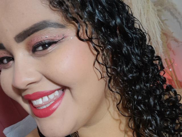 MelannieCarter's face with curly black hair, pink eyeshadow, glitter, red lipstick, and a smile showing white teeth. Her tan skin and slightly flushed cheeks are visible.