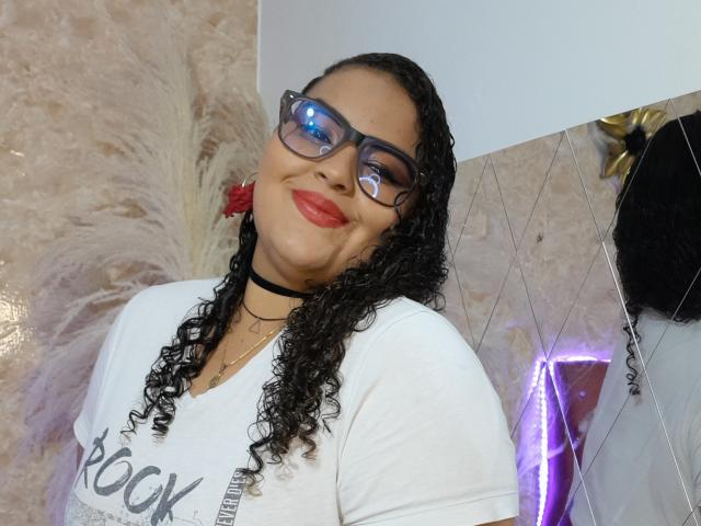 MelannieCarter with curly black hair, red lipstick, glasses, white shirt, black choker, smiling, large ass, medium tits, reflection in glasses, white and beige tiled wall, purple neon.
