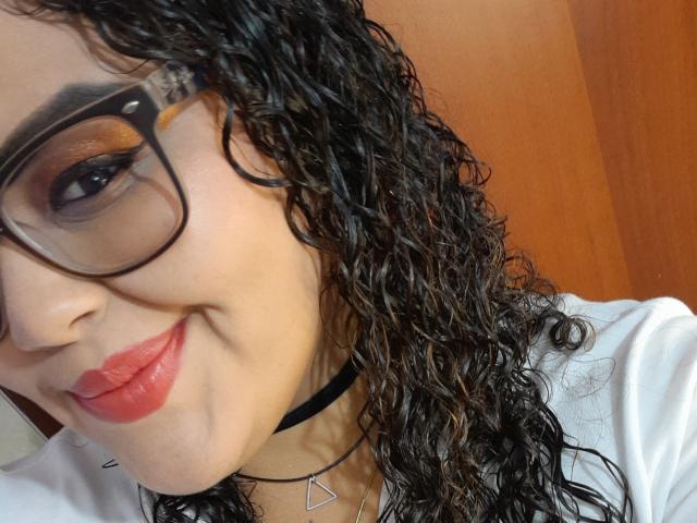 MelannieCarter with curly black hair, wearing glasses, red lipstick, black choker, white shirt, smiling, brown door in background. Boobs not visible. Face and upper body in focus.