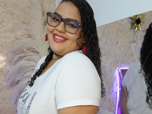 MelannieCarter with curly black hair, red earrings, glasses, white tee, red lipstick, smiling, showing her shiny teeth, and a confident look. Her tits are visible under her tee.