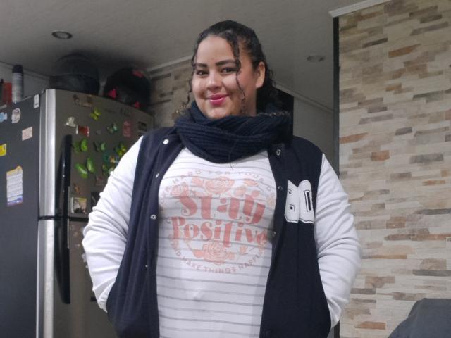 MelannieCarter has curly dark hair, a black scarf, white "Stay Positive" shirt, black vest, and pink lipstick, standing with hands in her pockets, smiling. Her tits are medium-sized.