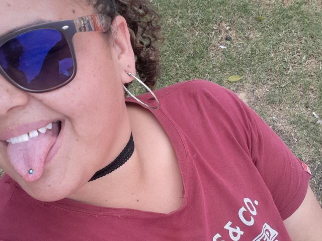 MelannieCarter with curly brown hair, wearing red shirt, black choker, and large hoop earrings, sticking out tongue with blue tongue piercing, and wearing reflective sunglasses.