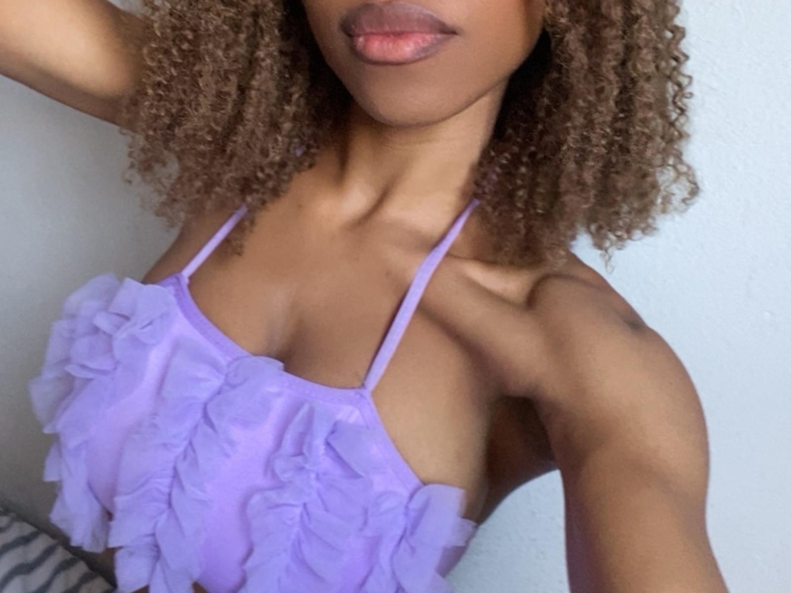 Ninah69 with curly brown hair, wearing a purple ruffled top, showing her small tits and hand on her neck, pink lips, and light brown skin.