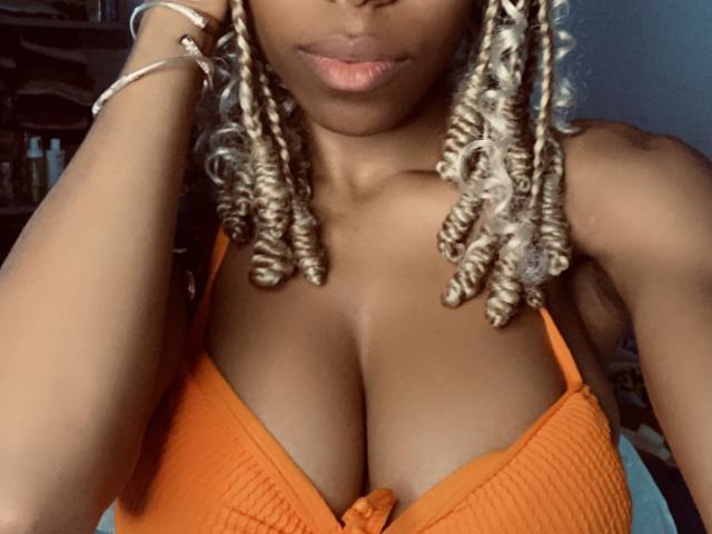 Ninah69 with dark brown skin, curly blonde-dipped braids, orange ribbed bikini top, showing her big boobs, slightly pouty lips, silver bracelet on right wrist, in a selfie.