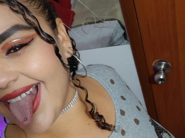 MelannieCarter's face, tongue out, showing a blue tongue piercing, wearing red lipstick, silver eyeshadow, curly brown hair, gray polka-dot top, and a pearl choker.