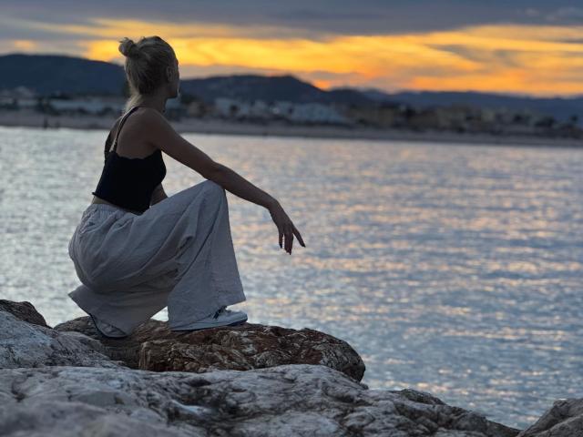 TinyJuly-hot with blonde bun, black tank top, and loose gray pants, squatting on rocky shore, looking at sunset over water. Her ass is slightly visible.