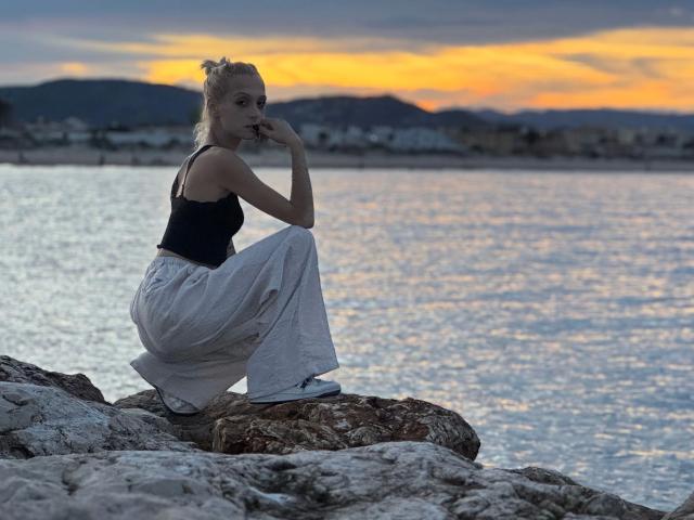 TinyJuly, con aspecto de estar agachada en una orilla rocosa, vestida con una camiseta negra y pantalones blancos holgados, con los dedos cerca de la boca, con el atardecer de fondo, el agua reflejando el cielo naranja, y el cabello rubio recogido en un moño..