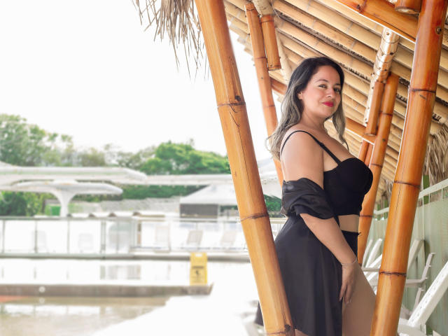 LolitaBusty with black lingerie, showing her tits and ass, standing against bamboo poles, looking at the camera, with a confident smile, and long dark hair.