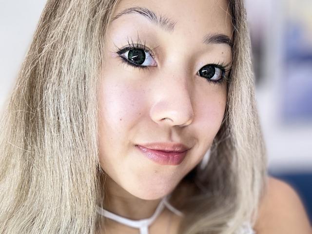 RenyLime's face with blonde hair, green eyes, and pink lips, wearing a white halter, showing a small mole on her left cheek. Her makeup includes long, thick eyelashes.