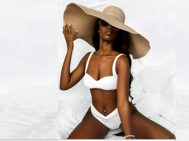 JessyBlacko with dark skin, white bikini, and big straw hat, sitting with legs apart, wearing sunglasses, showing small tits and a slim, toned ass, on white bed.