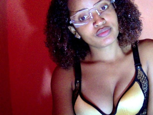Annaellah, avec ses cheveux bruns bouclés, ses lunettes et son anneau de nez, porte un soutien-gorge en dentelle jaune et noir, laissant entrevoir ses seins. L'arrière-plan est rouge. Elle sourit légèrement..