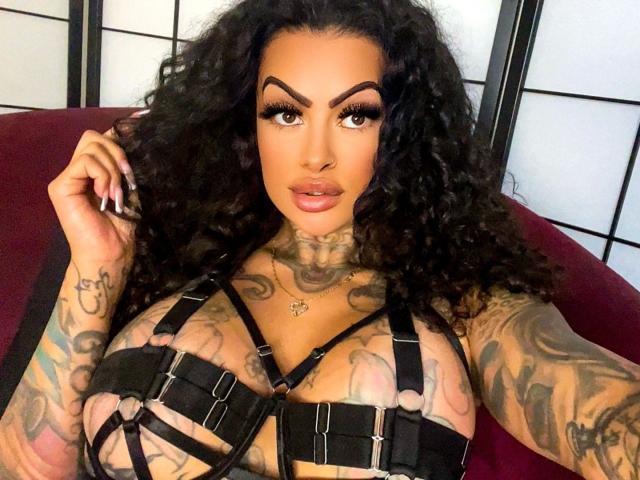 Leyla-del-Amor with curly black hair, dark eyebrows, full lips, tattoos on chest and arms, wearing black strappy lingerie, large tits, hand in hair, sitting on red chair.