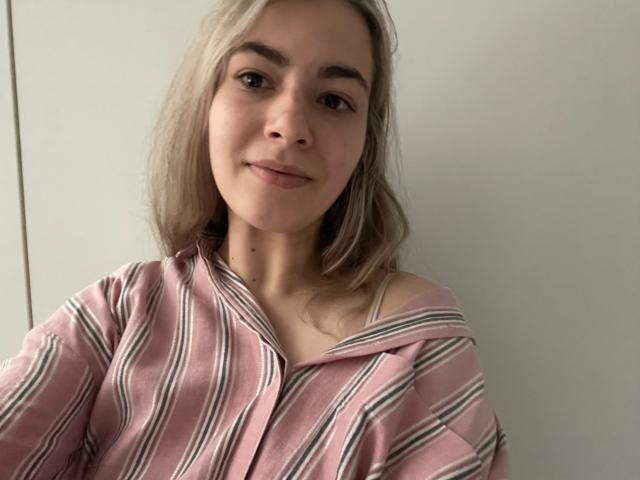 FancyRosie with light blonde hair, wearing a pink and white striped off-shoulder shirt, smiling softly at the camera. Her small tits are partially visible under the shirt.