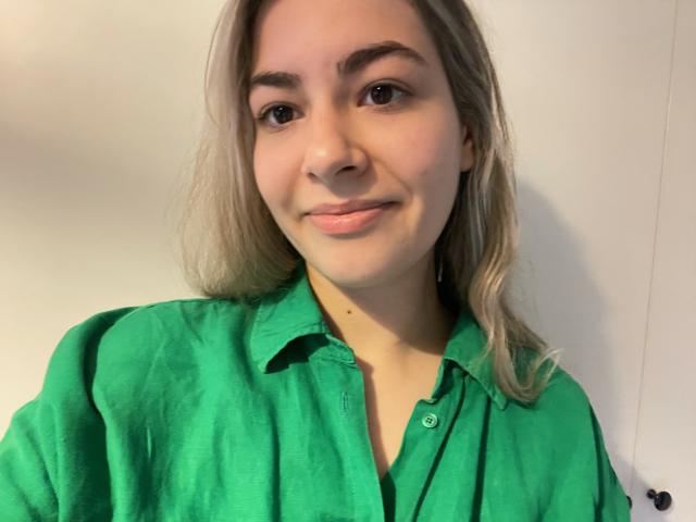 FancyRosie with blonde hair, green shirt, and slight smile, showing her cute face and small tits. Her brown eyes and light skin are visible. She's looking directly at the camera.