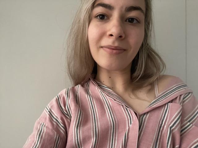 FancyRosie with blonde hair, wearing a pink and white striped off-shoulder shirt, smiling softly at the camera, showing a cute, subtle look with no makeup.