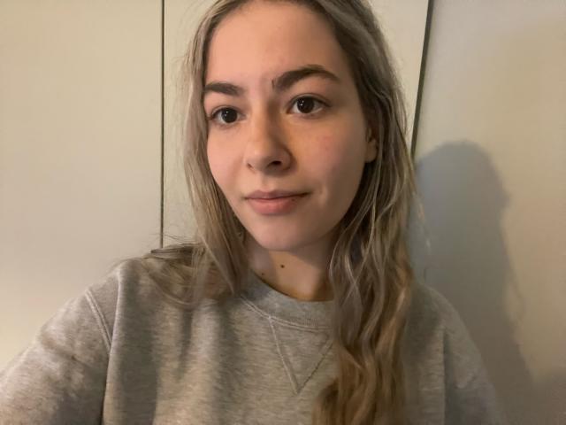 FancyRosie with long, wavy blonde hair, brown eyes, and a slight smile, wearing a grey sweatshirt, her cute face and small nose are visible.
