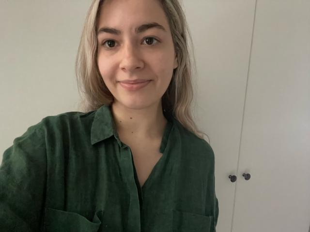 FancyRosie with long blonde hair, green button-up shirt, slight smile, and small moles on her face, showing upper body. Her tits are modest, and she's cute.