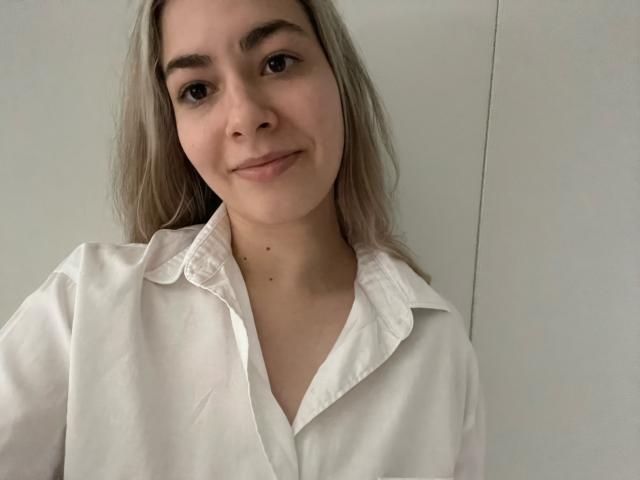 FancyRosie with light blonde hair, white button-up shirt, and subtle smile, showing small dark moles on her neck, soft brown eyes, and gentle expression.