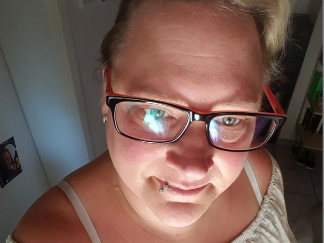 KleinerTeufel82-hot, chubby with pale skin, blonde hair, red-framed glasses, nose piercing, and lip piercing, wearing a white tank top, looking up at the camera.