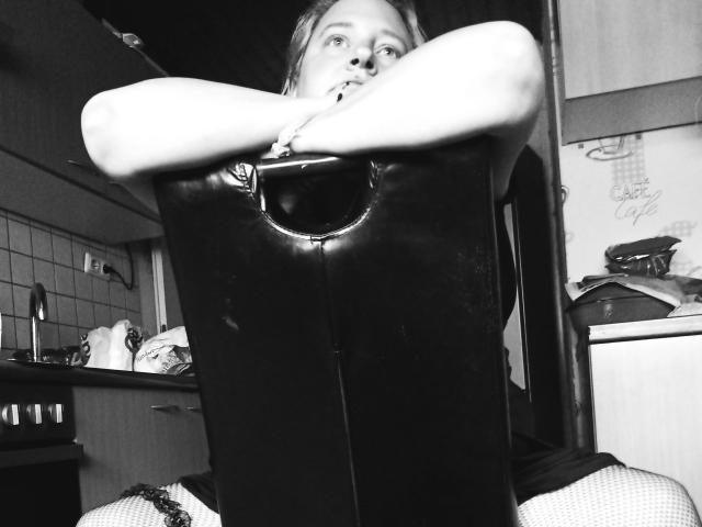 KleinerTeufel82-hot with arms crossed over black metal box, wearing fishnet stockings, looking up with a slightly open mouth, in a black-and-white photo.
