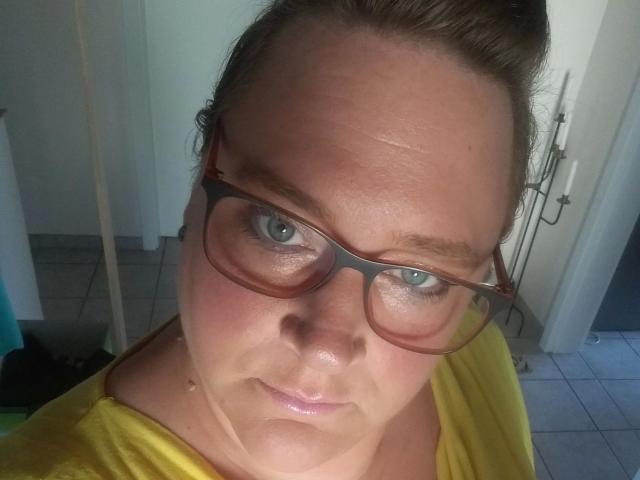 KleinerTeufel82-hot with glasses, blue eyes, and a yellow shirt, has a slightly shiny face, brown hair in a bun, and a neutral expression.