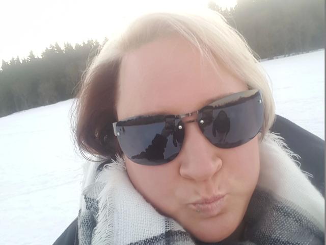 KleinerTeufel82-hot with blonde hair, black sunglasses, puffy nose, and a black-and-white checkered scarf, puckering their lips in a cold, snowy setting.