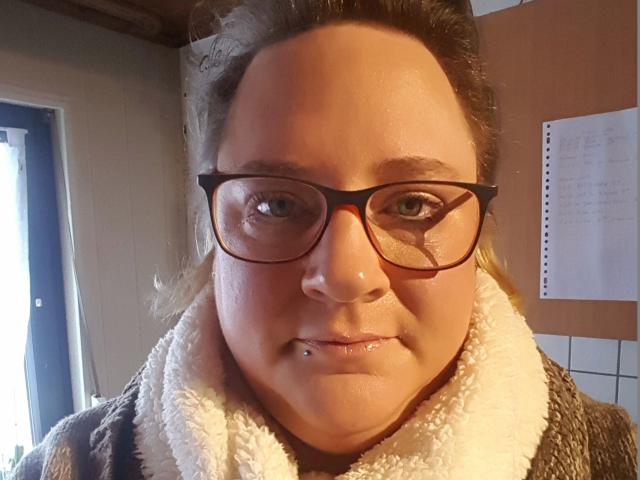 KleinerTeufel82-hot has a chubby face, glasses, and a nose piercing with a small silver ball. She's wearing a fluffy brown and white coat, smiling slightly.