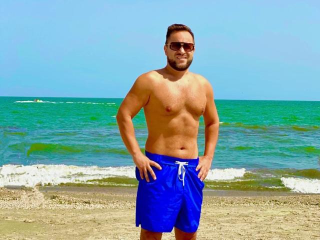 GabrielNastyGuy ist ein muskulöser, behaarter Mann ohne Hemd, der blaue Shorts, eine Sonnenbrille und ein Lächeln trägt und auf einem Strand mit grün-blauem Meer und weißen Wellen steht..