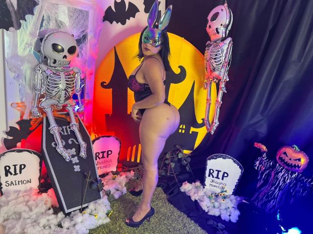ZahorySexy with curvy ass, large boobs, wearing a black lace bodysuit, metallic mask, and bunny ears, standing between two skeleton props with glowing eyes.