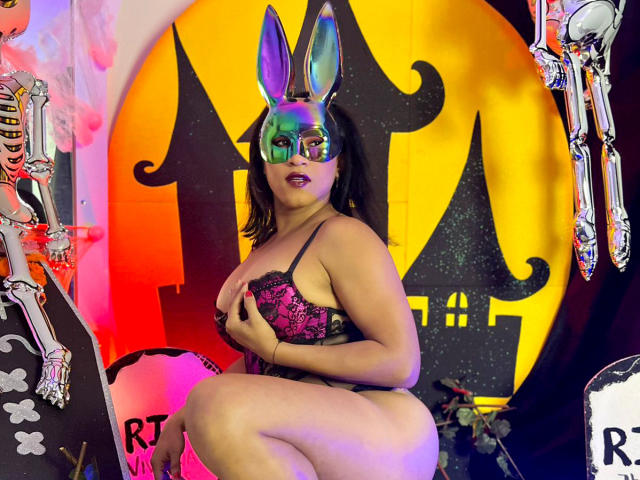 ZahorySexy with colorful bunny ears and glasses, wearing a pink lace bra, covering her tits with one hand, kneeling in a spooky Halloween-themed setup.