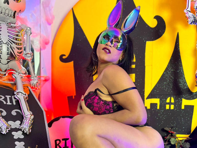 ZahorySexy, wearing a black lace bra and purple panties, crouches with shiny blue bunny ears and reflective mask, showing her tits and ass, with a skeleton on the left.