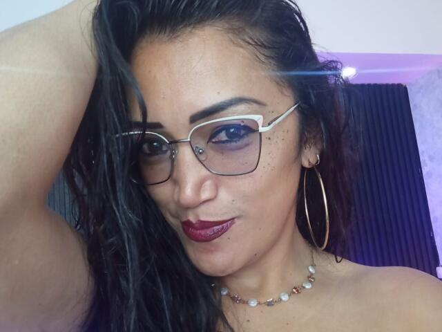 DayanaMarians69 met donker haar, een bril, rode lippenstift, oorringen en een parelketting, die haar arm omhoog steekt, waarbij haar schattige, sproetige gezicht en haar lichte glimlach te zien zijn..