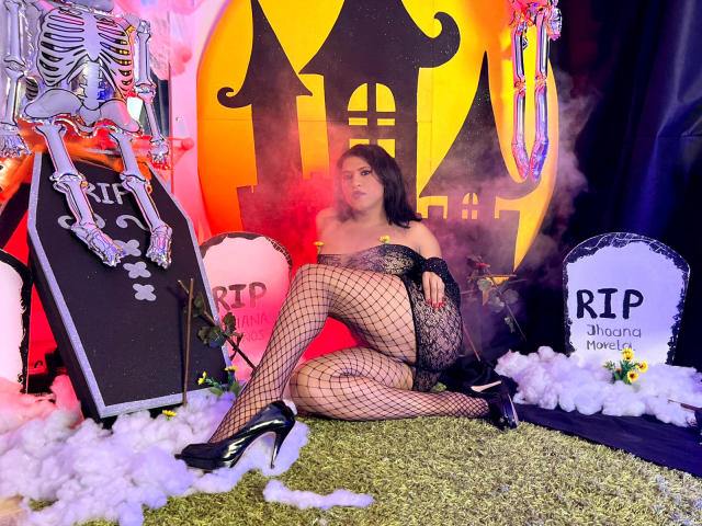 ZahorySexy with black lace lingerie, fishnet stockings, and black high heels, sitting on green carpet, showing her ass and tits, with a skeleton and RIP signs in background.
