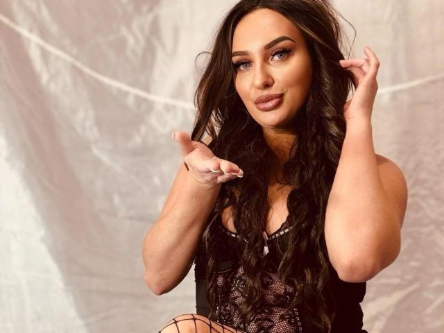 SweetSerena-hot with long wavy brown hair, wearing black lace lingerie, pointing with fingers up, showing off her tits and fishnet stockings, smiling with pink lipstick.